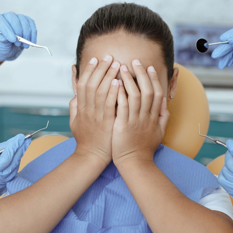 Multiracial doctors in rubber gloves scare girl with medical instruments. Mixed race little girl patient with napkin on chest in dental chair covers face with hands, close up, cropped, cropped